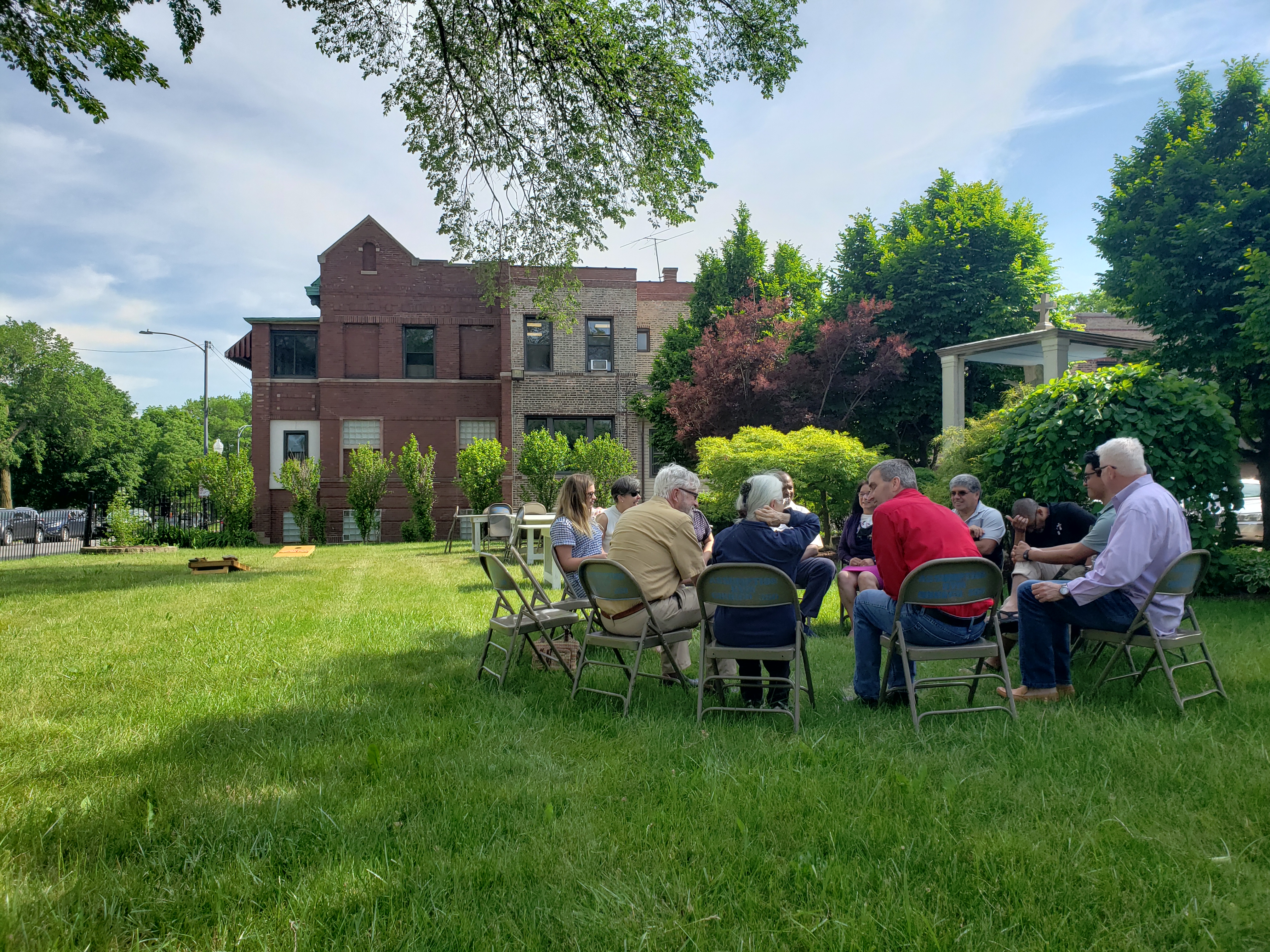 A group of people sit in a circle on folding chairs on a lawn in front of a house.